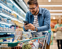 Ein Mann steht im Supermarkt mit Einkaufswagen und schaut auf sein Handy