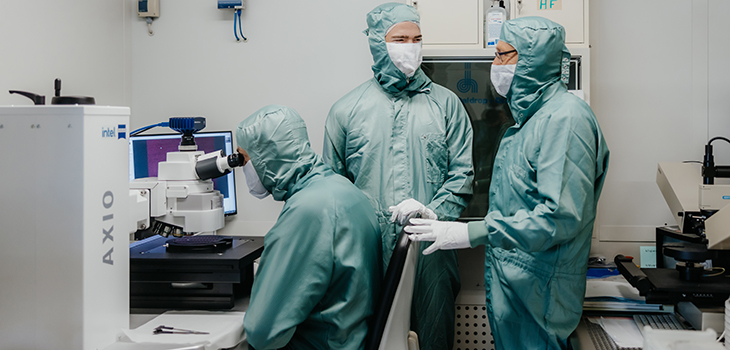 Rector Strackeljan talks to two trainees in the cleanroom, one is sitting at the microscope